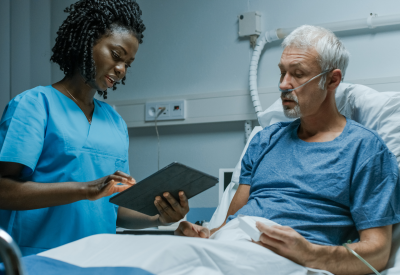 Une infirmière munie d'une tablette électronique parle à un patient âgé à l'hôpital.