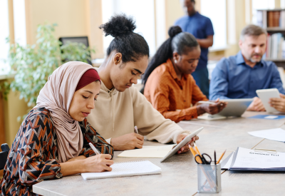 Groupe de personnes d'origines ethniques diverses prenant des cours de langue pour les immigrants, assis à une table et effectuant des tâches à l'aide de tablettes numériques.